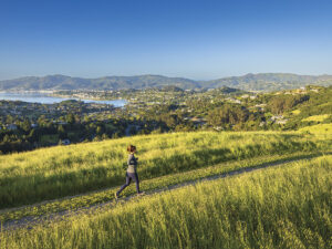 woman jogging on trail in grassy area with mountains and water in background with blue sky in Tiburon