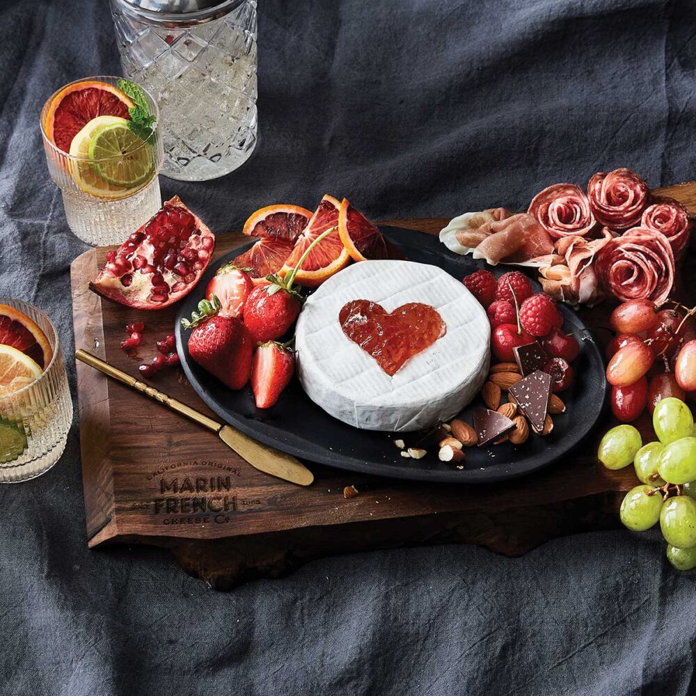 Marin French Cheese Valentine Board showing cheese with heart shaped jam, fruit, on wood board displayed with cocktails on gray tablecloth for Cheese Trail article