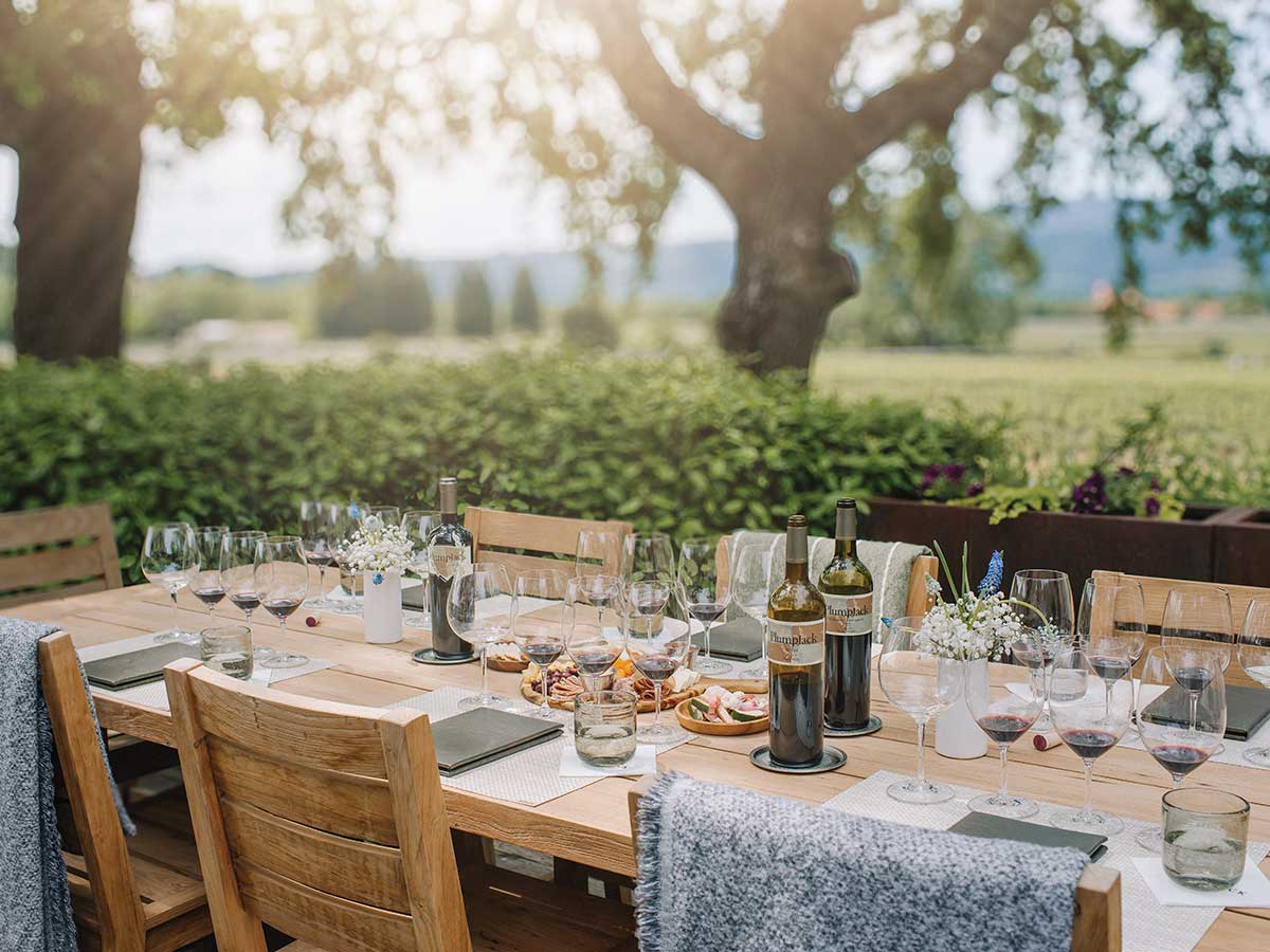outdoor wooden table set in green garden with plates, glasses, placemats and bottles of PlumpJack Winery wine in Oakville