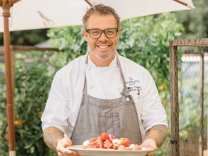 Chef Patrick Prager at Silverado Resort in white chef shirt, gray apron and glasses holding a plate with brightly colored red tomatoes, standing outside under white umbrella, smiling