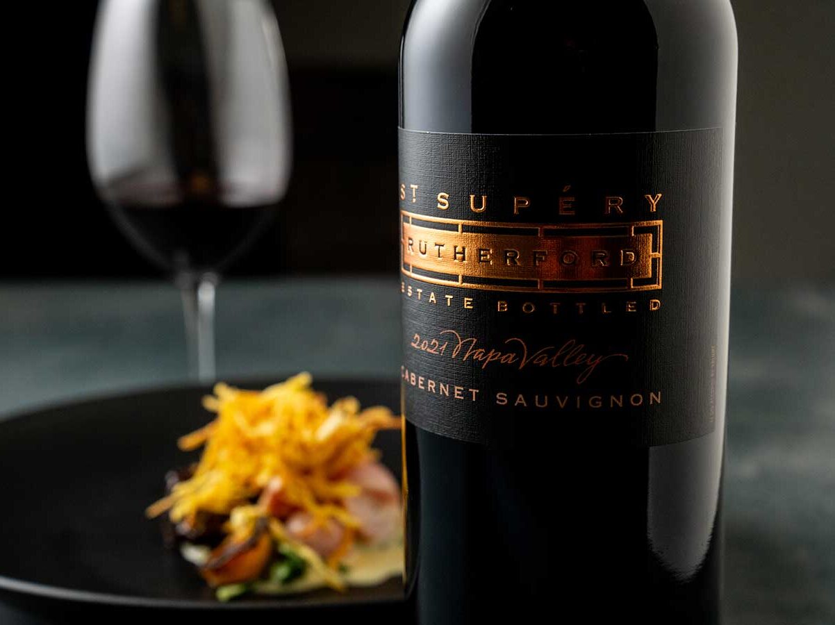 Bottle of St. Supéry wine next to plate of food with glass of red wine on table