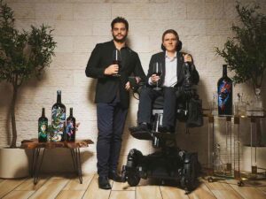 Blake Barrios of Rebellium dressed in black suit holding a glass of red wine with Hawken Miller in gray suit and white shirt holding a glass of red wine and bottle of wine in chair against stone wall at Thyme Well Spent Catering “Kitchen Collective”